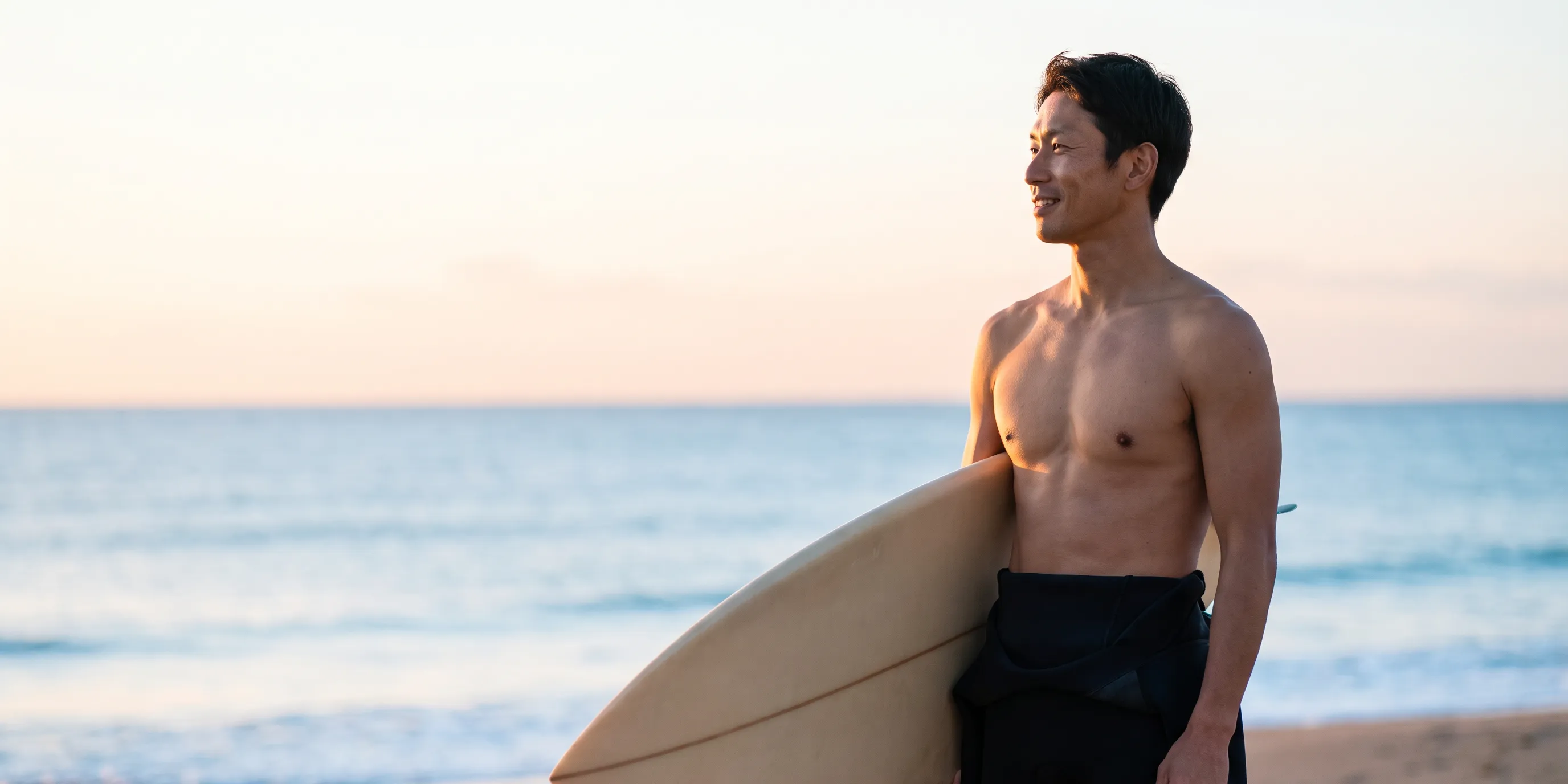 朝の海辺で、サーフボードを抱えて朝日を見る男性