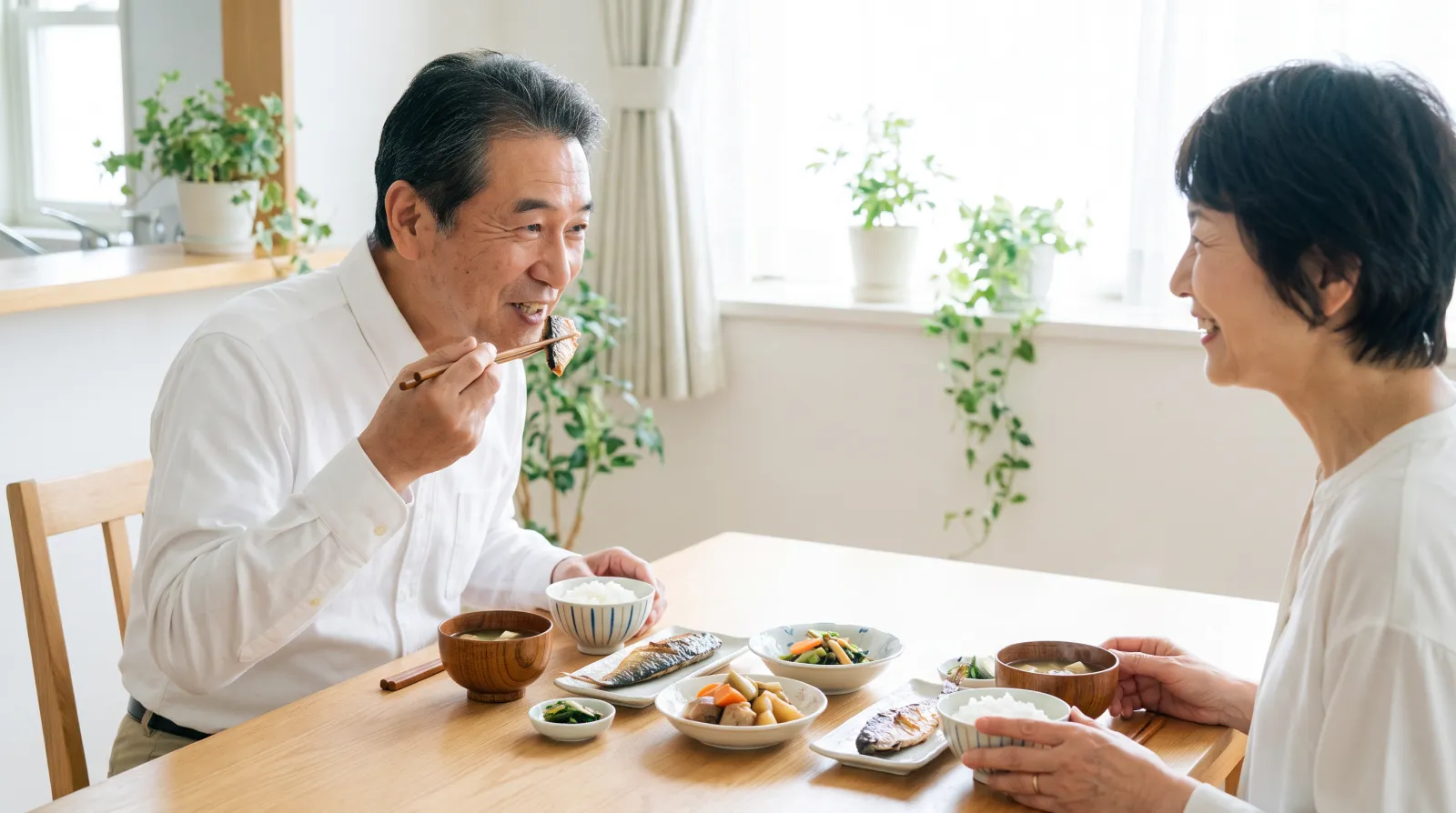 明るい食卓で、60代の夫婦が向かい合って和食を楽しんでいる。夫は箸で料理を口に運びながら笑顔