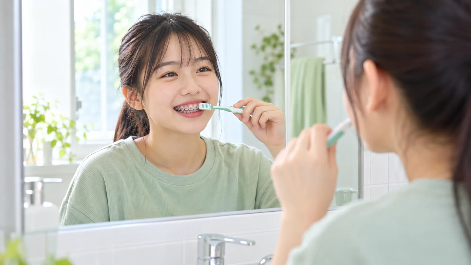 明るい洗面所で、装置をつけた高校生の女の子が鏡の前で笑顔で歯をみがいている