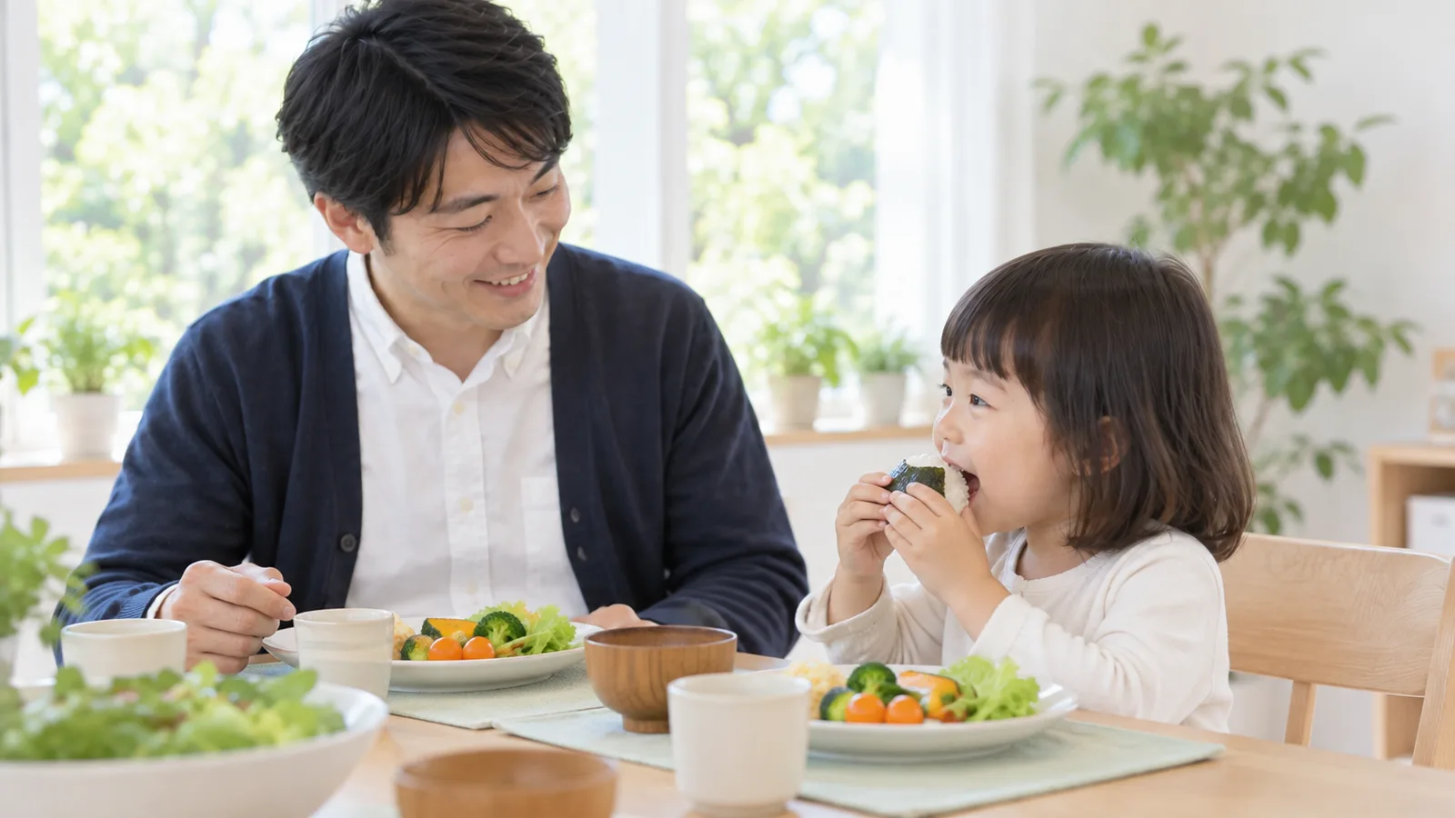 食卓で、幼い女の子が海苔で巻いたおにぎりを両手で持ち、父親がそばで見守っている