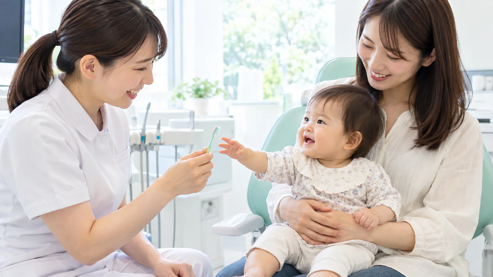 明るい診療室で、母親のひざに座った1歳の赤ちゃんが、女性の歯科医師の持つ小さな歯ブラシに手を伸ばしている