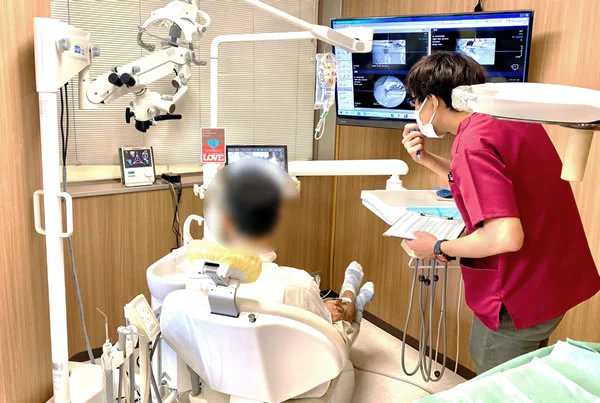 診療室で、歯科医師が壁のモニターに映したレントゲンとCTの画像を指しながら、診療台に座った患者さんに説明している（患者さんの顔はぼかし）