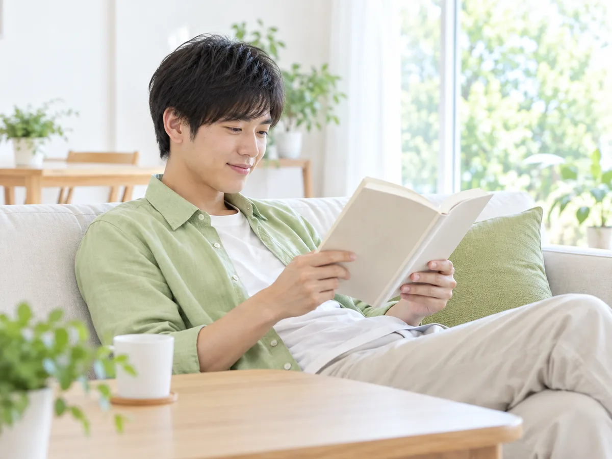 自宅のソファで、大学生の男性がクッションにもたれて本を読みながら休んでいる