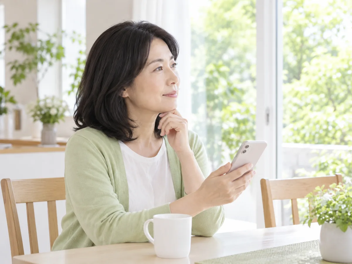 自宅のダイニングで、40代の女性がスマートフォンを手に、少し考えている