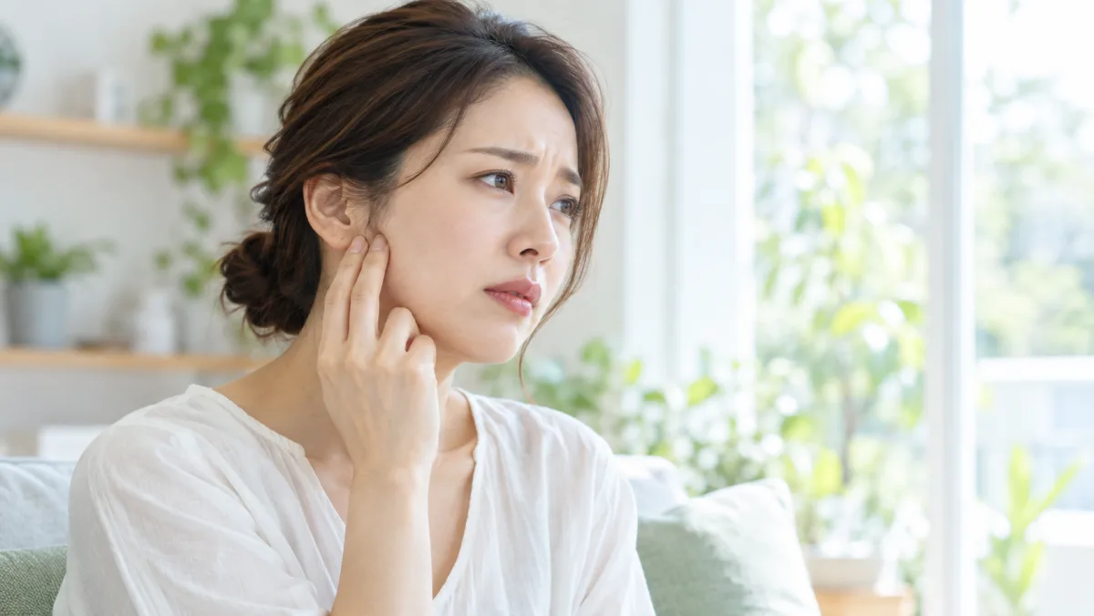 自宅の窓辺で、30代の女性が耳の前のあごの関節のあたりを指先で押さえ、少し不安そうにしている