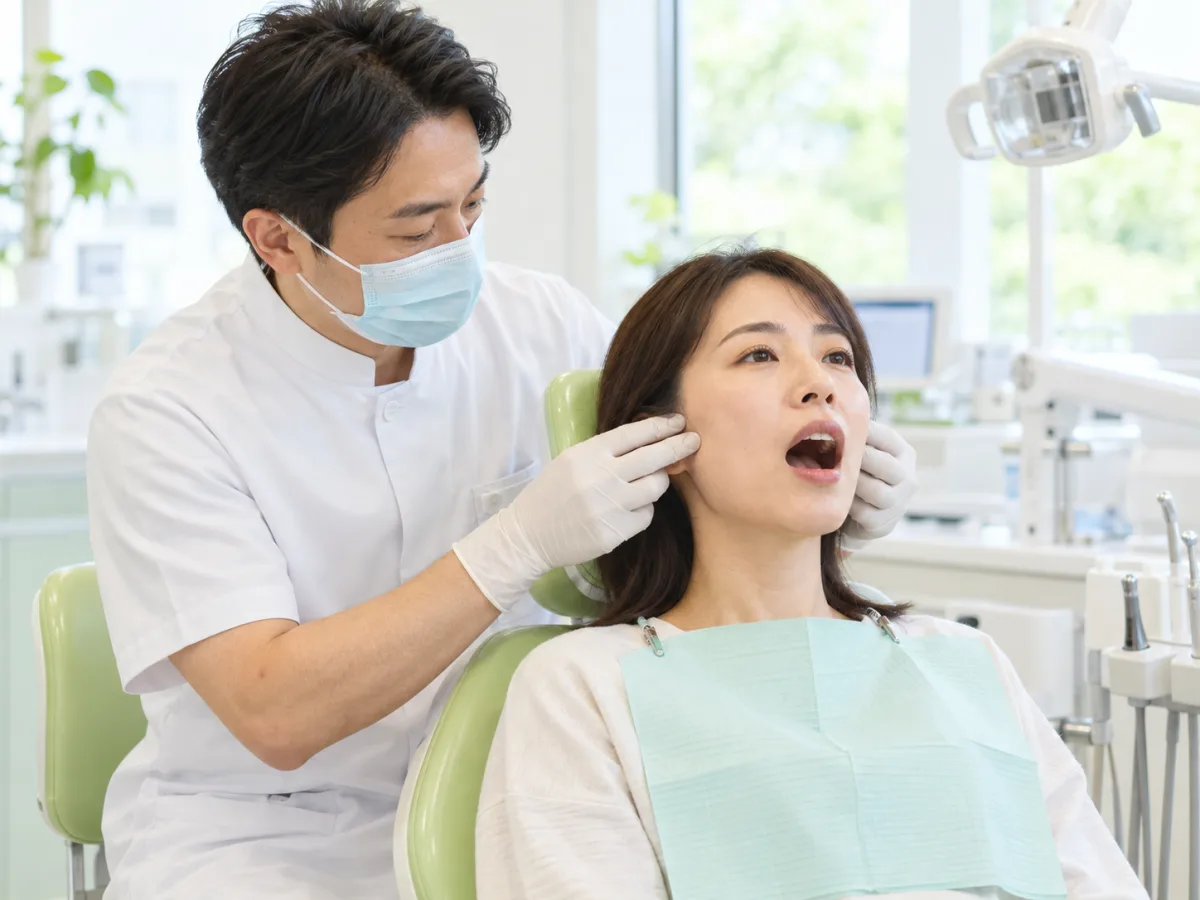 診療台で、歯科医師が女性の耳の前のあごの関節に指先を当て、口を開けたときの動きを確かめている