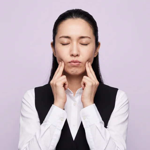 目を閉じて歯を食いしばり、あごの筋肉を指で押さえる女性